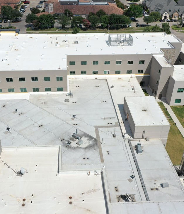 Aerial view of a large white-roofed industrial or commercial building complex with surrounding greenery and roads.