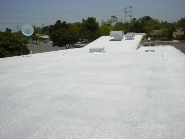 Flat white rooftop with ventilation units, surrounded by trees and distant power lines under a clear blue sky.