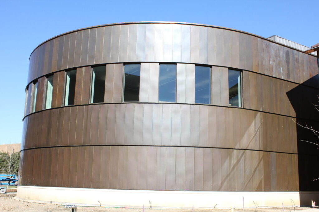 A modern, circular building with vertical metal paneling and multiple windows under a clear blue sky.
