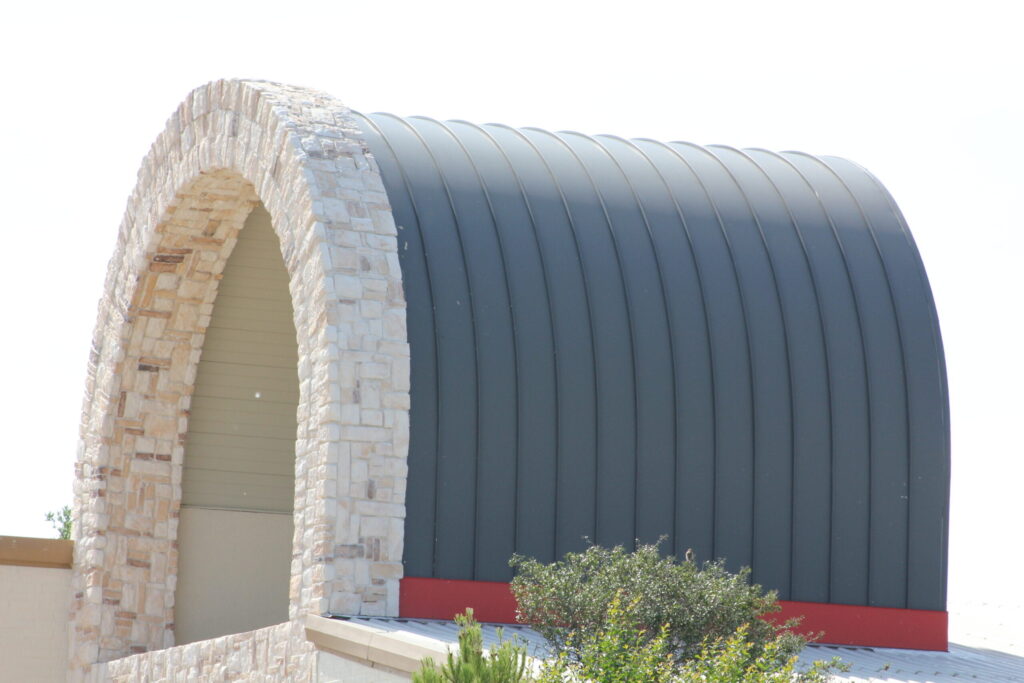 A building with a rounded, stone arch and a curved, ribbed metal roof against a clear sky.