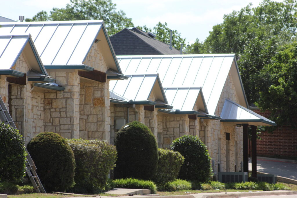 Stone building with a metal roof, surrounded by trimmed bushes and trees, under a clear sky.