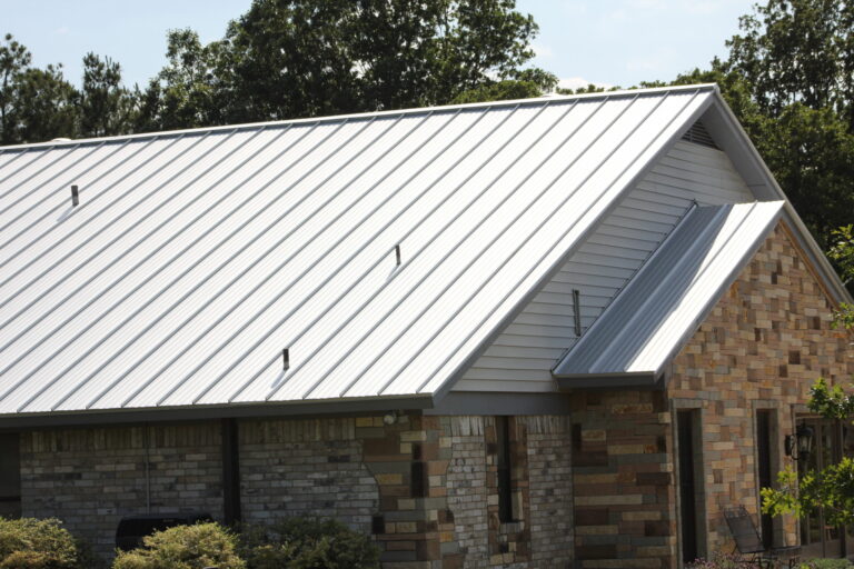 A house with a sloped metal roof and brick exterior, surrounded by trees and bushes.