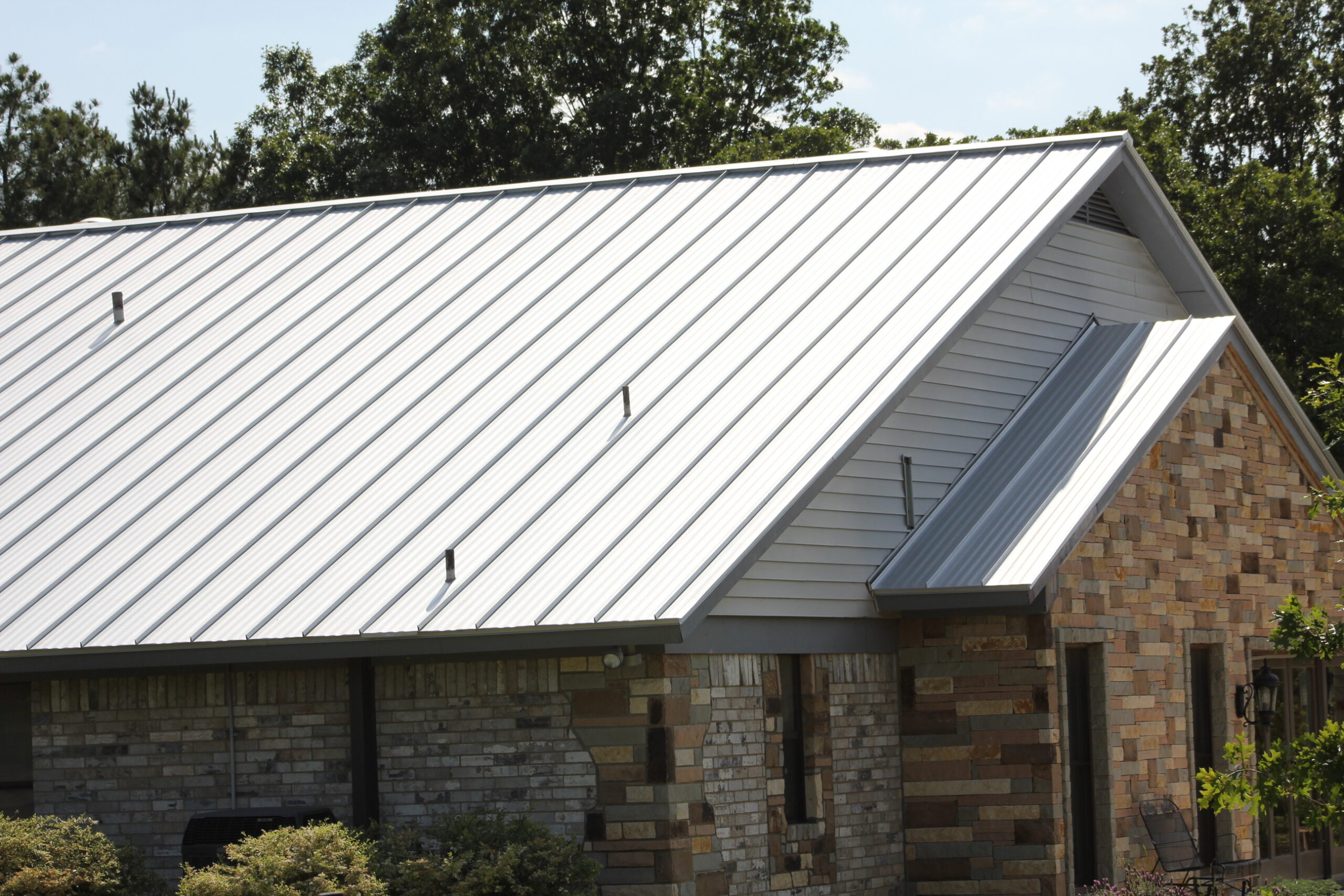 A house with a sloped metal roof and brick exterior, surrounded by trees and bushes.