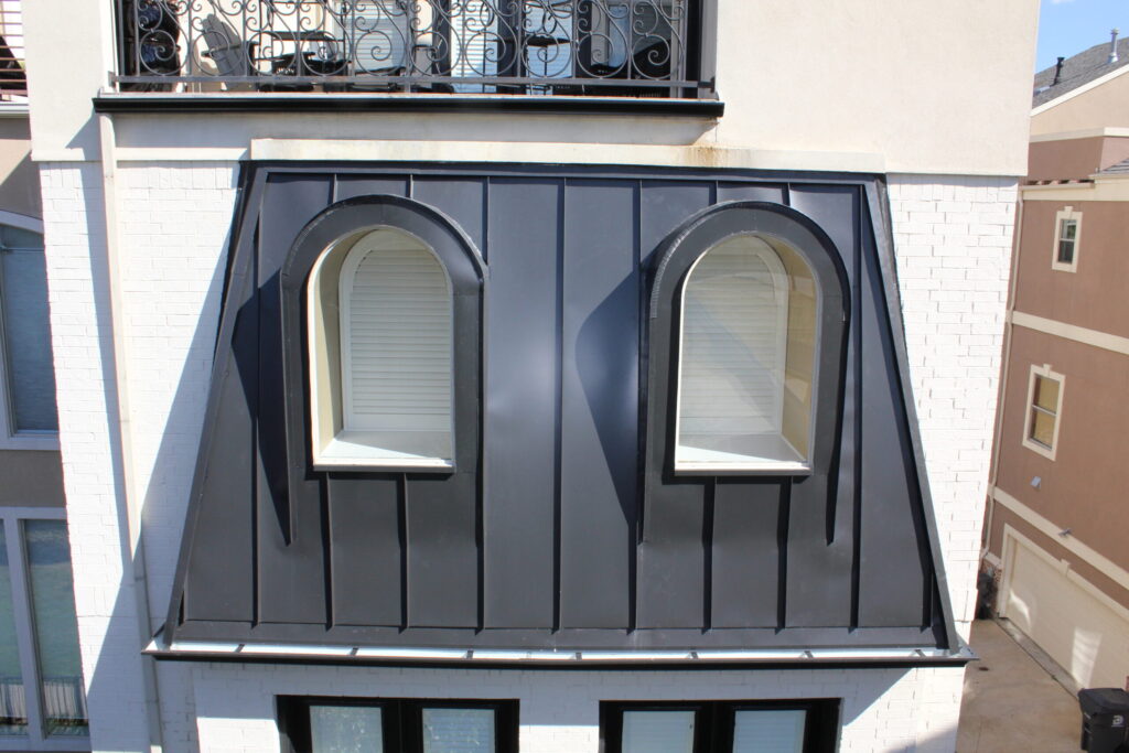 Black mansard roof with two arched dormer windows, set against a white brick wall.
