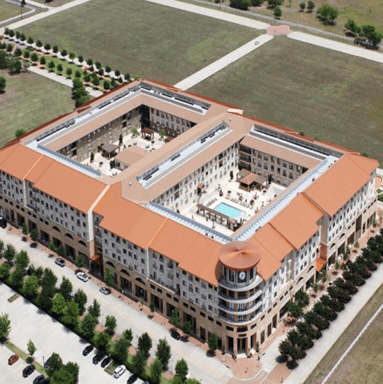 Aerial view of a U-shaped building with a courtyard, pool, and orange roof, surrounded by parking and greenery.