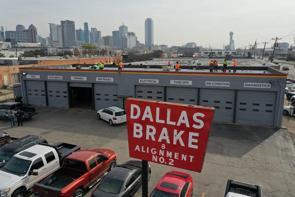 An aerial view captures the Dallas Brake & Alignment garage with workers diligently tending to commercial roofing tasks. The impressive city skyline serves as a stunning backdrop.