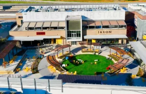 Aerial view of an outdoor dining area with a circular lawn, surrounded by seating and stylish restaurants like Jaxon and Shoulas. The scene is complemented by the pristine commercial roofing that shelters these vibrant eateries.
